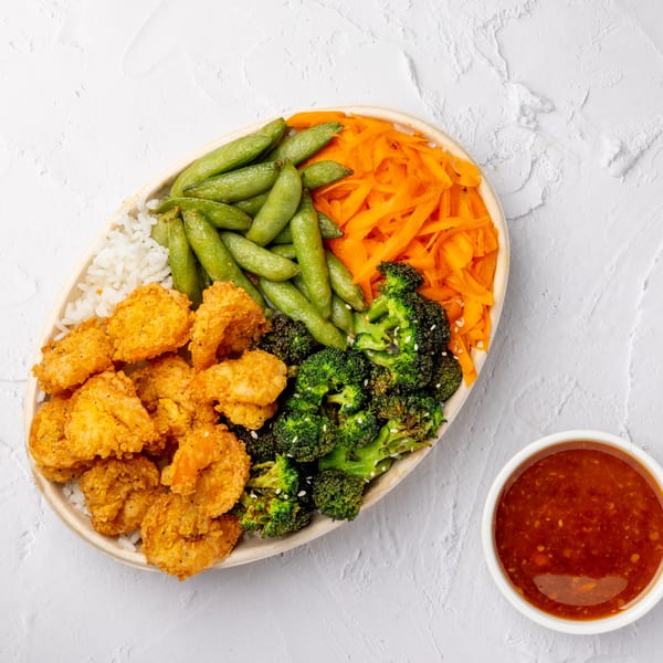 Bowl of fried chicken, rice, edamame, shredded carrots, roasted broccoli, and dipping sauce on a white textured surface.