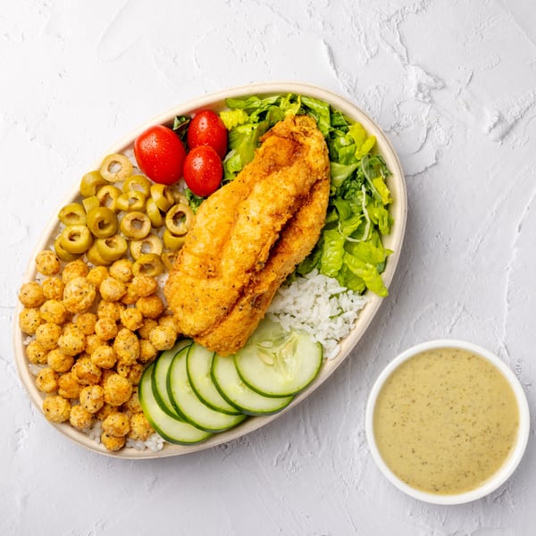 Mediterranean rice bowl with crispy chicken, roasted chickpeas, lettuce, tomatoes, olives, cucumber, and creamy herb sauce.
